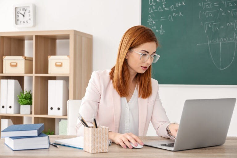 Beautiful,Young,Teacher,With,Laptop,Sitting,At,Table,In,Classroom